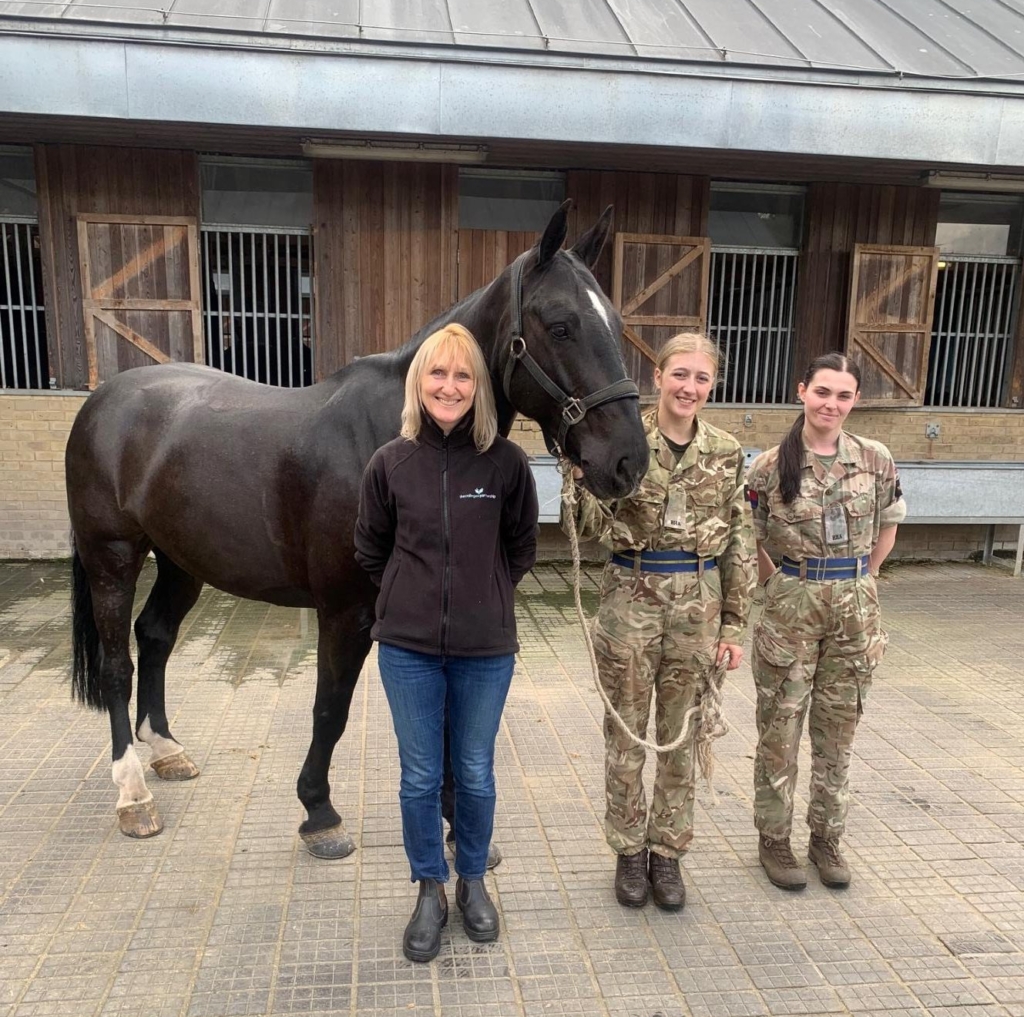 2 young female Army Apprentices in uniform stand next to black horse with civilian woman in front of stables